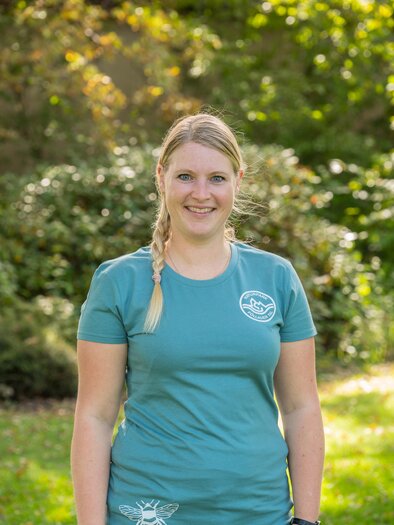 A woman with blonde hair stands in a green garden. She is wearing a blue T-shirt and is smiling warmly at the camera. | © Naturpark Pöllauer Tal