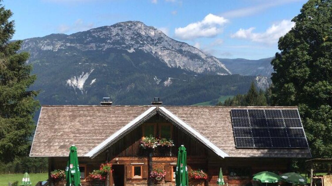 Ein traditionelles Holzhaus mit Solarpanelen in einer malerischen Berglandschaft. Es gibt grüne Sonnenschirme und Blumen auf der Veranda. | © Neureit-Alm