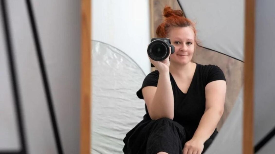 Eine Person mit rotem Haar sitzt im Trendoutfit und hält eine Kamera in der Hand. Im Hintergrund sind reflektierende Studiosoftboxen zu sehen. | © Nikolett Rosta