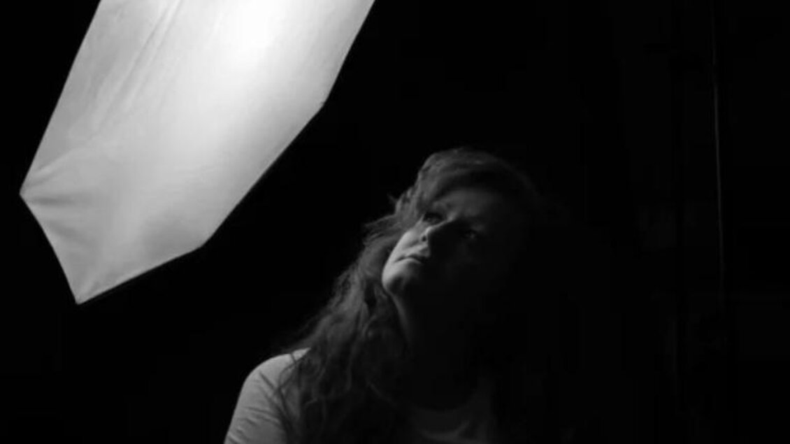 Eine Frau mit lockigem Haar posiert unter einem Studiolicht. Das Bild ist in Schwarzweiß gehalten und erzeugt eine dramatische Atmosphäre. | © Nikolett Rosta