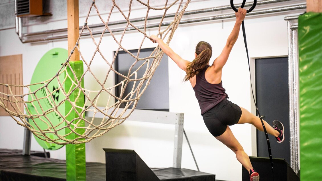 Frau klettert an einem Ninja Warrior Parcours in der Ninjabox Graz, während sie sich an einem Seil festhält und über ein Netz schwingt. | © Ninjabox