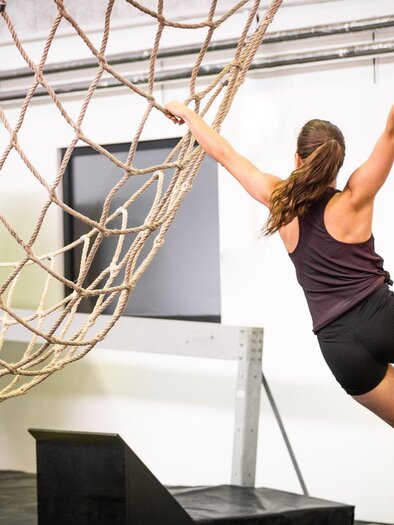 Frau klettert an einem Ninja Warrior Parcours in der Ninjabox Graz, während sie sich an einem Seil festhält und über ein Netz schwingt. | © Ninjabox