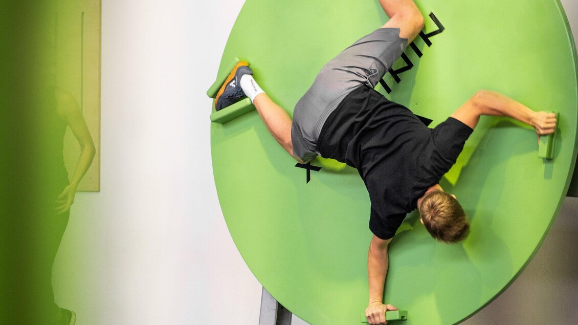 Junger Mann klettert an einer Trainingsscheibe  in der Ninjabox Graz, indem er sich mit speziellen Griffen festhält und die Balance hält. | © Ninjabox