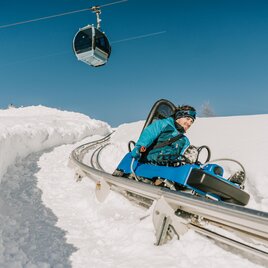 A person is riding a chain carousel on a snow sledding track. In the background, a cable car can be seen against a blue sky. | © TMG Turracher Höhe Marketing GmbH