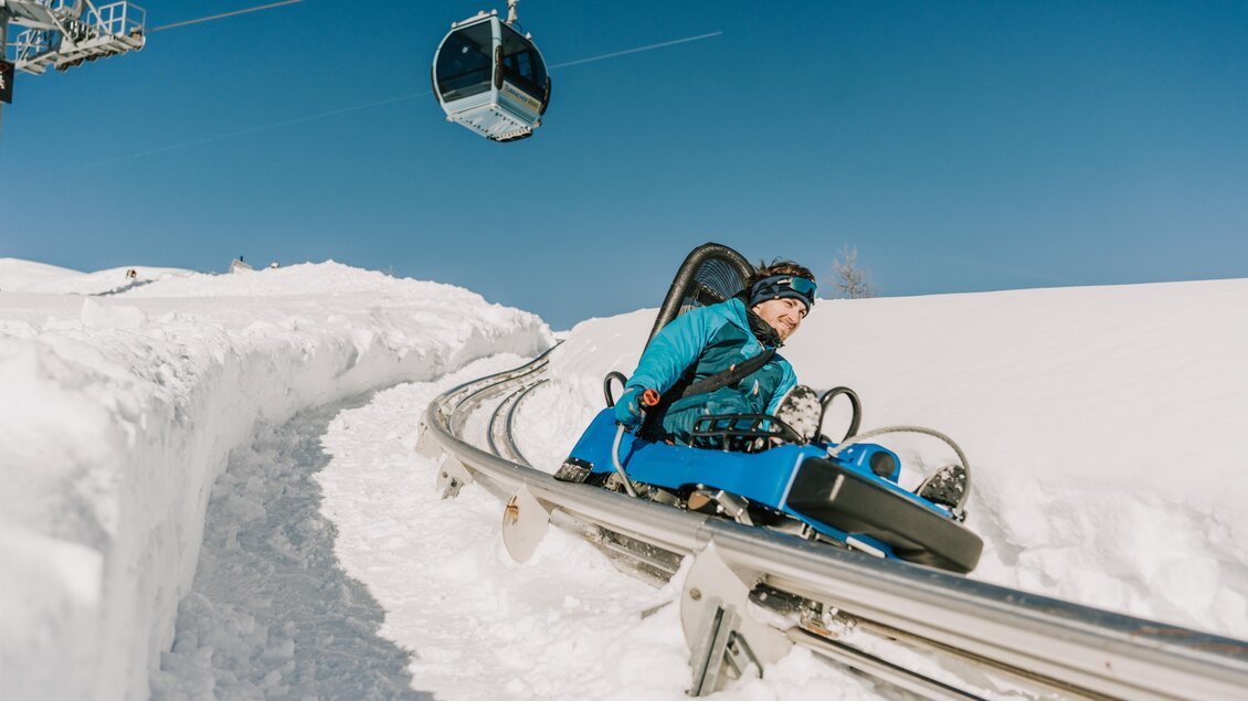 Eine Person fährt mit einem Kettenkarussell auf einer Schnee-Rodelbahn. Im Hintergrund ist eine Seilbahn vor blauem Himmel zu sehen. | © TMG Turracher Höhe Marketing GmbH