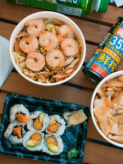 Ein Tablett mit verschiedenen asiatischen Gerichten, darunter Nudeln mit Garnelen, gebratenes Hähnchen und Sushi. Dazu ein Getränk in einer bunten Dose. | © Noodle King – Michael Choup