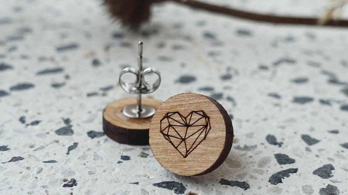 Ein handgefertigter Ohrring aus Holz mit einem eingravierten Herzdesign liegt auf einer steinernen Oberfläche. Im Hintergrund sind zarte Pflanzen sichtbar.