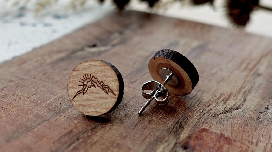 Zwei Holzohrringe mit einem Bergmotiv und einer Sonne sind auf einem Holzuntergrund platziert. Die Ohrringe haben einen natürlichen und rustikalen Look.