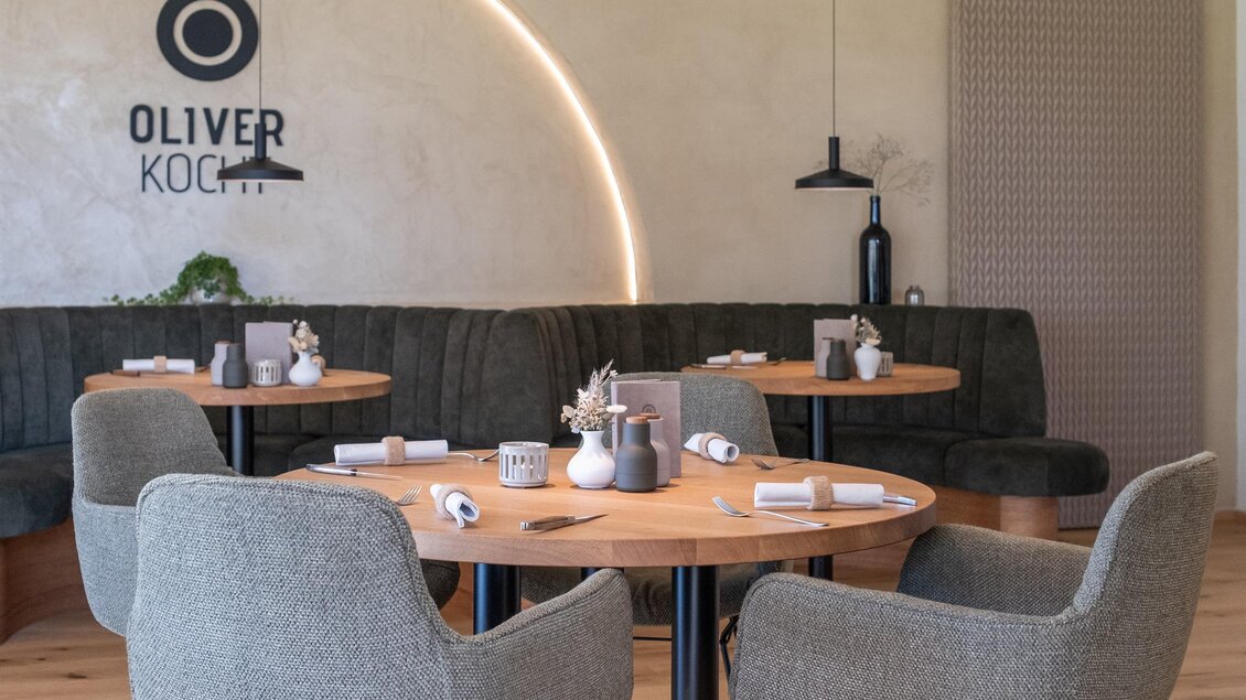 Ein modern eingerichteter Essbereich mit einem runden Tisch und beigen Stühlen. An der Wand befindet sich das Logo "OLIVER KOCHT". | © Oliver kocht