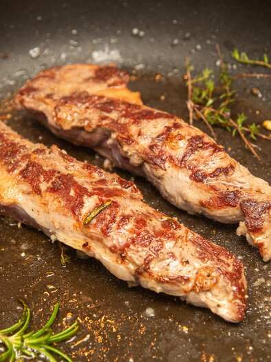 Two grilled pieces of meat in a pan, surrounded by herbs and garlic. The surface of the steaks is nicely browned and appetizing. | © AdobeStock_688130530