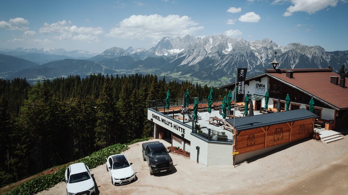Eine Berglandschaft mit schneebedeckten Gipfeln und einer klaren Blauen Himmel. Im Vordergrund steht eine Almhütte mit Terrassen und mehreren Autos. | © OWH