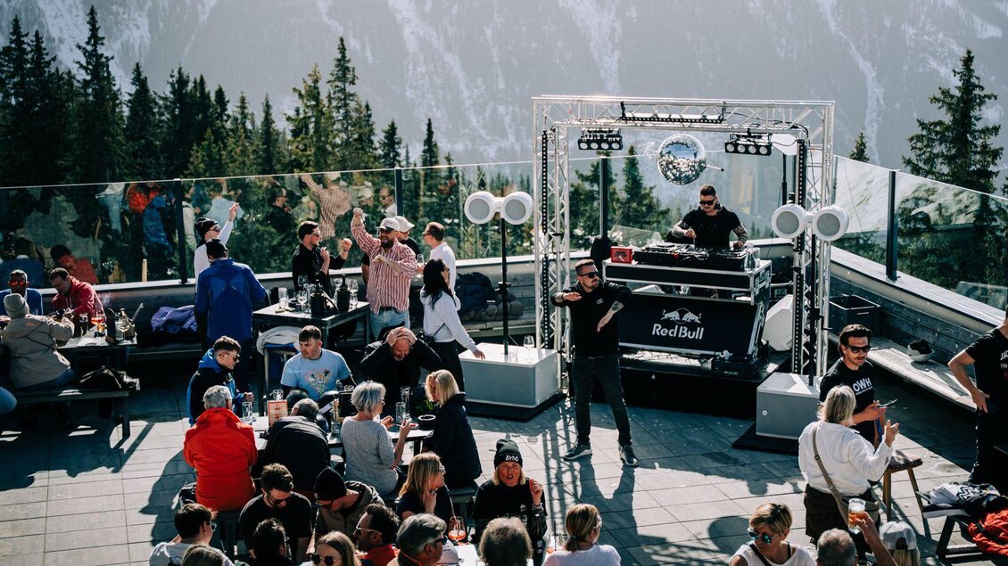 Eine lebhafte Alm-Lounge mit Menschen, die tanzen und eine DJ-Performance genießen. Im Hintergrund sind hohe Berge und ein sonniger Himmel zu sehen.