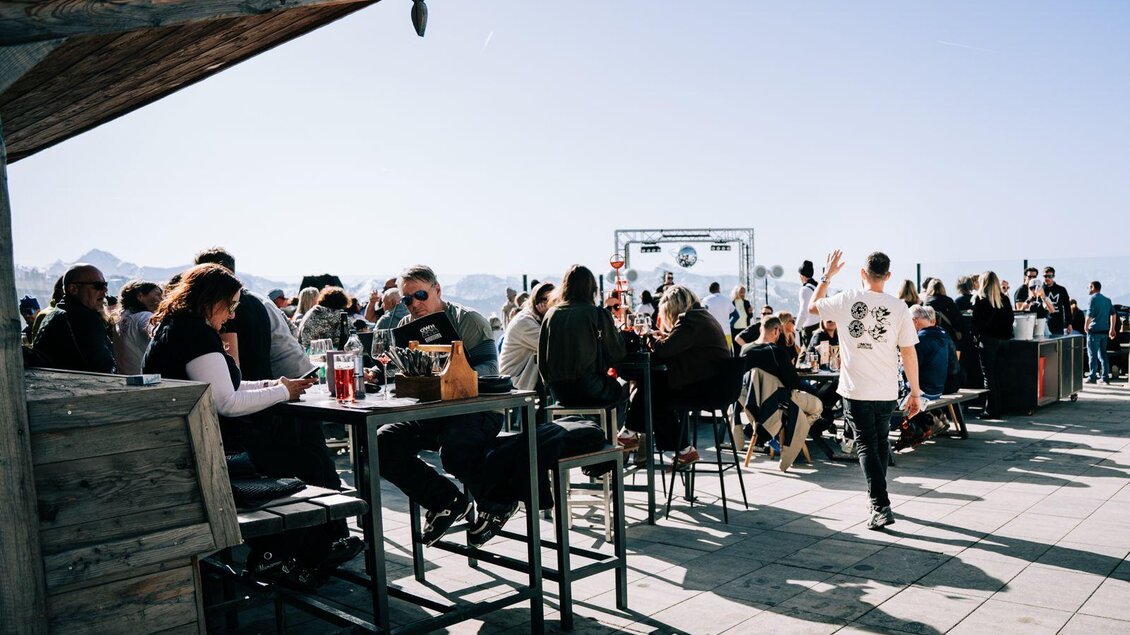 Eine belebte Terrasse mit vielen Leuten, die bei schönem Wetter gemeinsam Zeit verbringen. Im Hintergrund sind Berge und eine Bühne zu sehen.