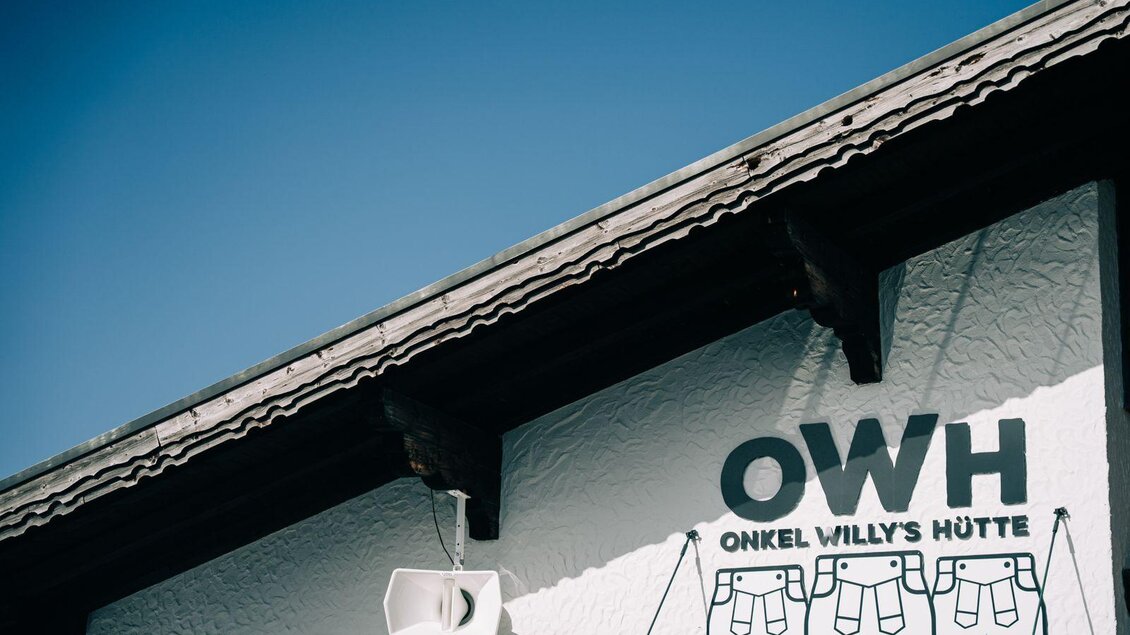Ein Teil eines Gebäudes mit einem weißen Wanddesign. Der Schriftzug "Onkel Willy's Hütte" und ein Lautsprecher sind sichtbar.