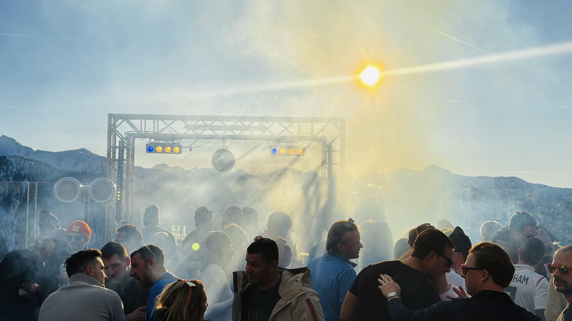 Eine lebhafte Party mit vielen Menschen und rauchiger Atmosphäre. Im Hintergrund ist eine Bühne mit Lichtstrahlen sichtbar.