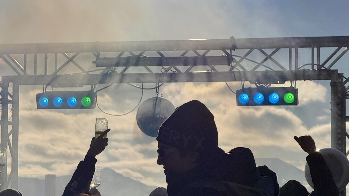 Eine lebhafte Menschenmenge feiert vor einer Bühne mit bunten Lichtern. Der Himmel ist blau und die Sonne scheint hell.