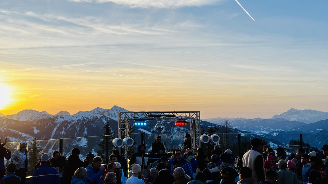 Eine lebendige Veranstaltung in den Bergen mit einer Menschenmenge vor einer Bühne. Im Hintergrund ist ein schöner Sonnenuntergang und eine klare Himmel zu sehen.