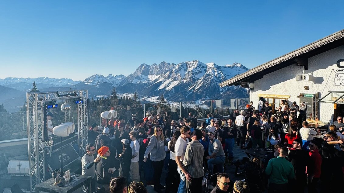 Eine lebhafte Veranstaltung im Freien mit vielen Menschen. Im Hintergrund sind beeindruckende Berge und ein strahlend blauer Himmel zu sehen.