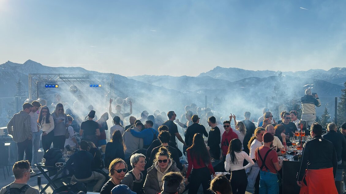 Eine lebhafte Veranstaltung im Freien mit vielen Menschen in der Sonne. Im Hintergrund sind Berge und Rauch zu sehen.