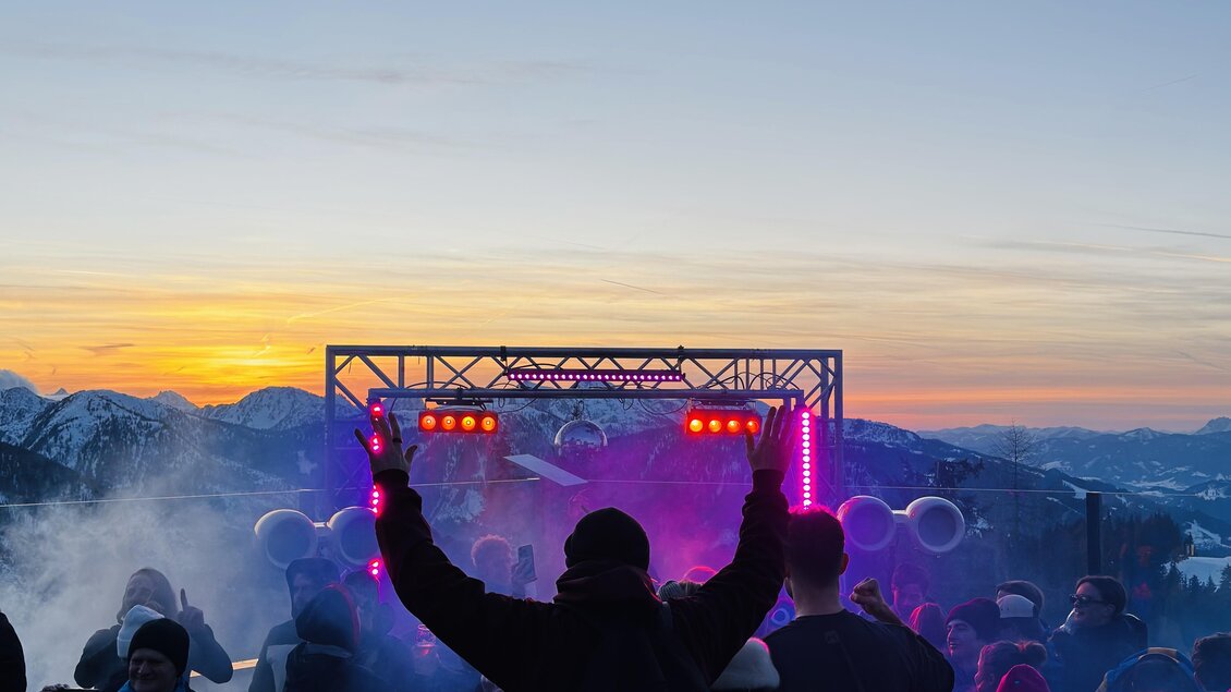 Eine lebendige Party in den Bergen bei Sonnenuntergang. Menschen feiern vor einer Bühne mit bunten Lichtern und Nebel.