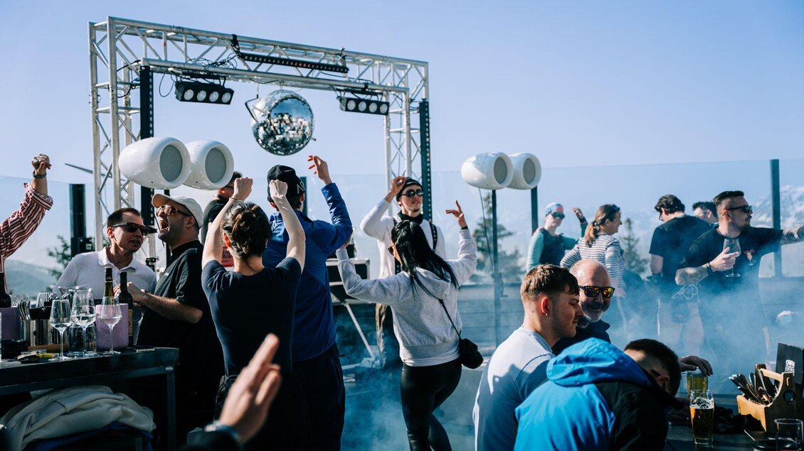 Eine lebhafte Party mit Menschen, die tanzen und feiern. Eine Discokugel und Musikboxen sorgen für eine festliche Atmosphäre im Freien.