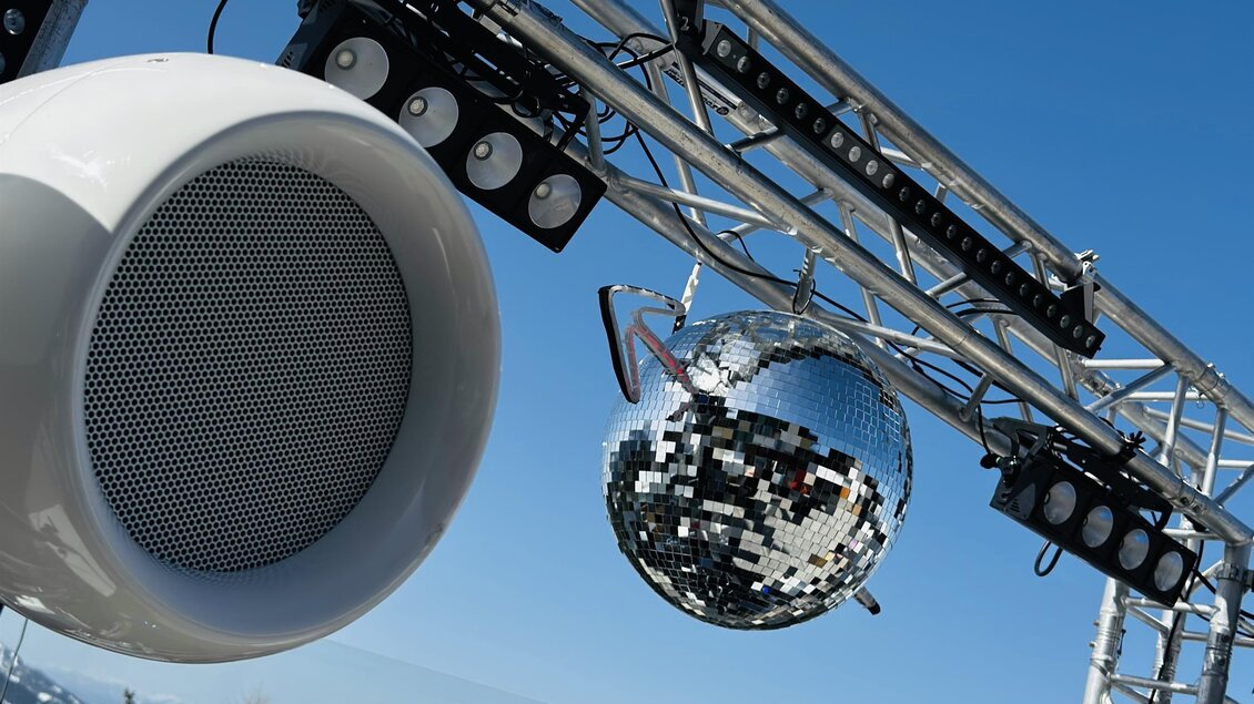 Eine Veranstaltungstechnik-Anlage mit Lautsprechern und einer Discokugel vor einer wunderschönen Berglandschaft. Der Himmel ist klar und blau.