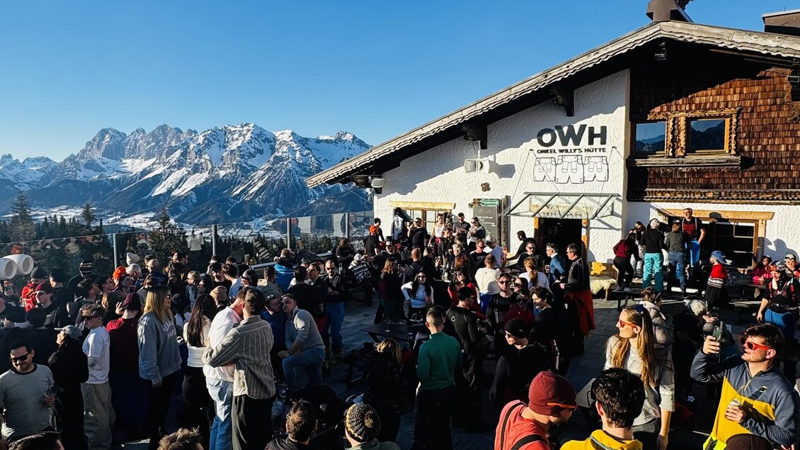 Ein belebter Ort in den Bergen mit vielen Menschen, die sich versammeln. Im Hintergrund sind schneebedeckte Berge und blauer Himmel zu sehen.
