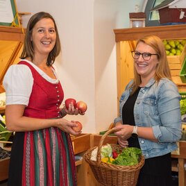 Zwei Frauen stehen in einem Obst- und Gemüsegeschäft. Sie lächeln und halten frisches Obst und Gemüse in den Händen. | © Bauernladen Pöllau