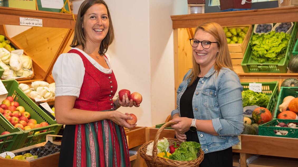 Zwei Frauen stehen in einem Obst- und Gemüsegeschäft. Sie lächeln und halten frisches Obst und Gemüse in den Händen. | © Bauernladen Pöllau