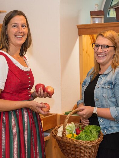 Zwei Frauen stehen in einem Obst- und Gemüsegeschäft. Sie lächeln und halten frisches Obst und Gemüse in den Händen. | © Bauernladen Pöllau