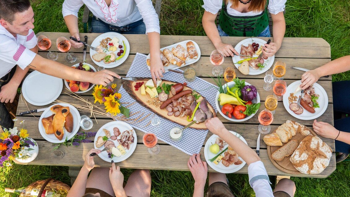 Eine festliche Tafel im Freien mit verschiedenen Gerichten, darunter Wurst, Salat und Brot. Mehrere Personen genießen das Essen zusammen in einer sommerlichen Atmosphäre. | © Bauernladen Pöllau