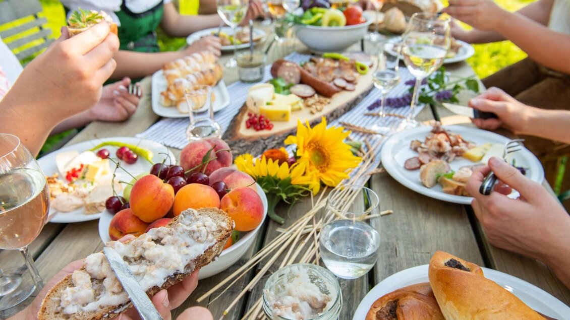 Ein schöner Tisch im Freien, gedeckt mit einer Vielzahl von Speisen und Getränken. Menschen genießen ein Festmahl mit frischem Obst, Brot und dekorativen Blumen. | © Bauernladen Pöllau