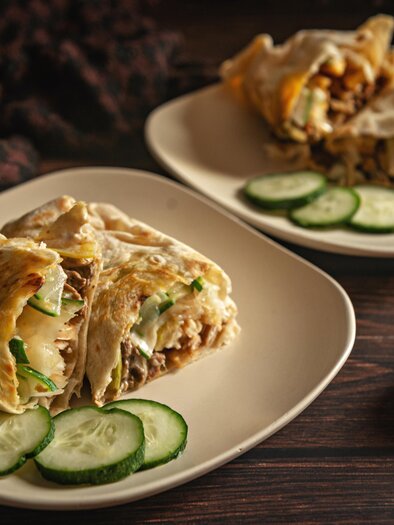 Ein Teller mit gefüllten Wraps, serviert mit Gurkenscheiben. Im Hintergrund liegen Knoblauch und Zwiebeln. | © pexels-dee-dave