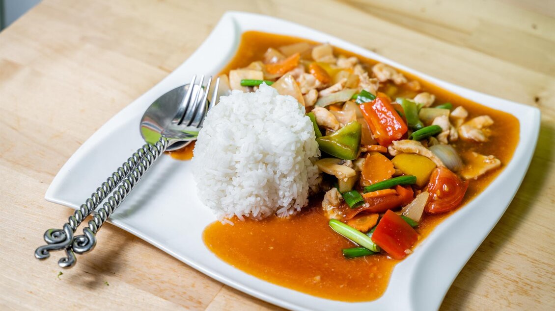 Ein Teller mit gebratenem Hühnchen und buntem Gemüse in einer Sauce, serviert mit einer Portion Reis. Das Gericht liegt auf einem weißen Teller auf einem Holztisch. | © Stephan Friesinger