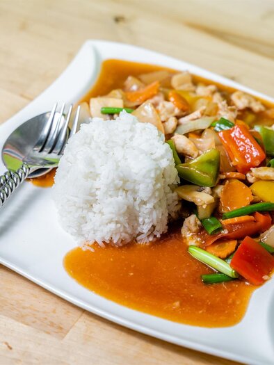 Ein Teller mit gebratenem Hühnchen und buntem Gemüse in einer Sauce, serviert mit einer Portion Reis. Das Gericht liegt auf einem weißen Teller auf einem Holztisch. | © Stephan Friesinger