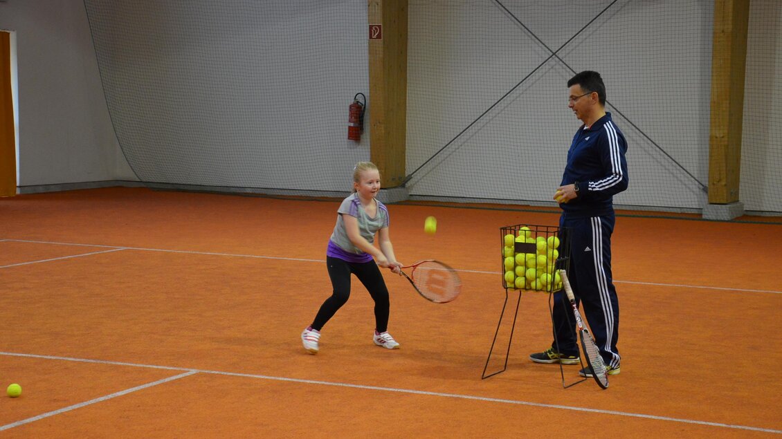 Ein Mädchen spielt Tennis in einer Halle und schlägt einen Ball. Ein Trainer steht neben einem Korb mit Tennisbällen und beobachtet sie. | © POSS Freizeithalle