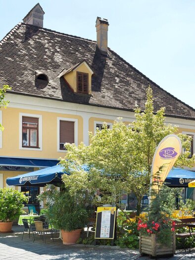 A charming restaurant on a sunny street, surrounded by colorful plants and patio furniture. The yellow facade of the building exudes coziness. | © TV Region Graz - René Vidalli