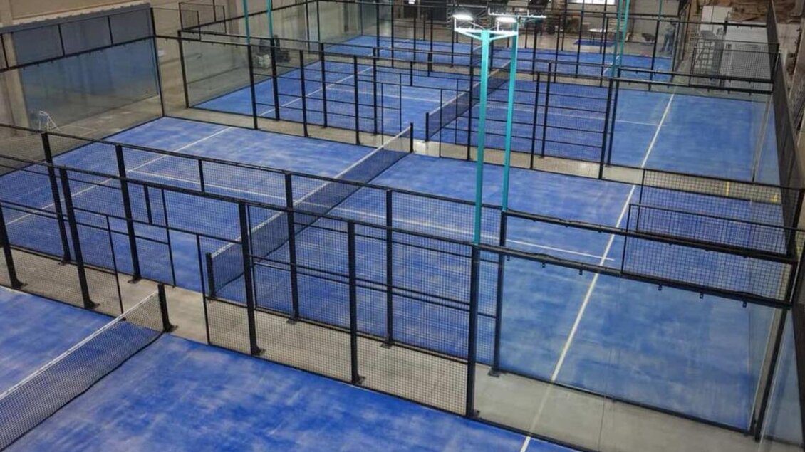 Ein Innenbereich mit mehreren Padelplätzen, die durch schwarze Gitterwände abgetrennt sind. Der Boden ist in einem auffälligen Blau gehalten.