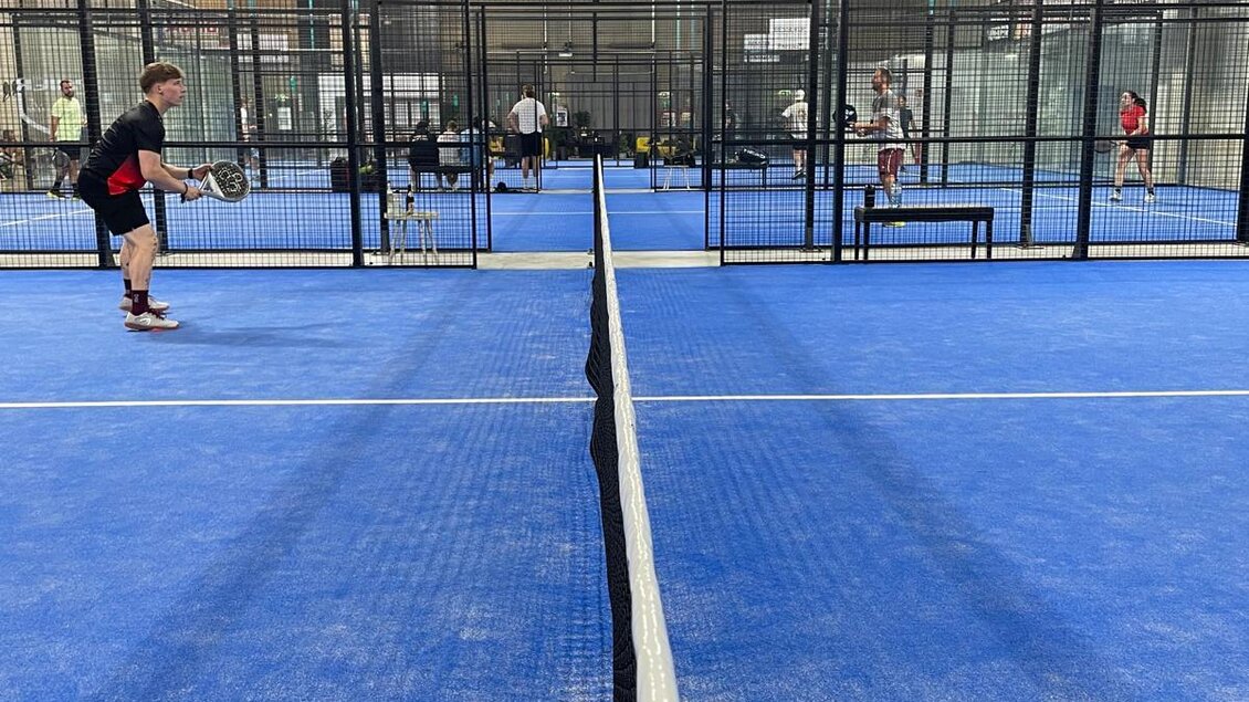 Ein gut beleuchteter Indoor-Tennisplatz mit blauen Spielfeldern. Spieler üben und spielen miteinander auf jeweils getrennten Plätzen.