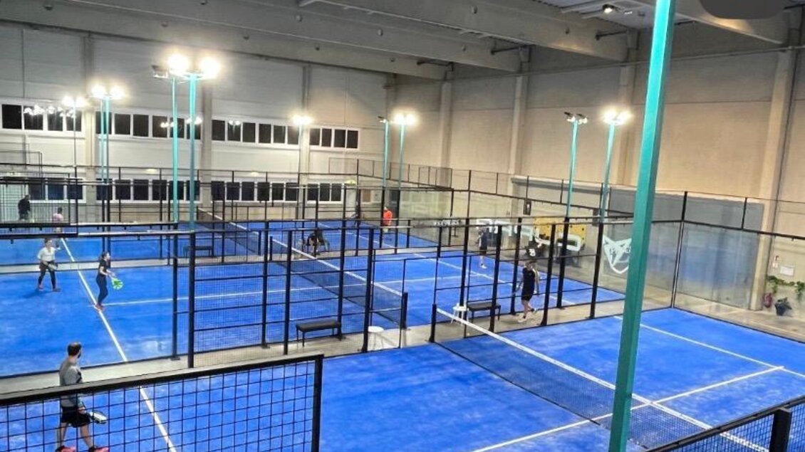 Ein moderner Sportplatz mit blauen Spielfeldern und Netzen. Mehrere Personen spielen aktiv Tennis oder Padel in einer gut beleuchteten Halle.