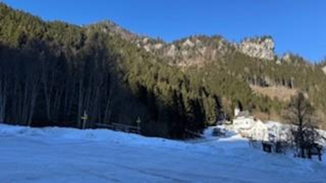 Ein winterlicher Landschaftsausblick mit schneebedecktem Boden und hohen Bergen im Hintergrund. Ein kleines Haus ist zwischen den Bäumen zu sehen. | © Hanna Giraudo