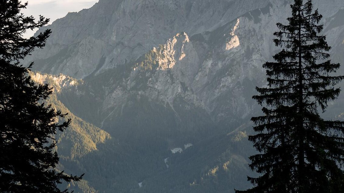 Eine beeindruckende Berglandschaft mit hohen Gipfeln und Wäldern im Vordergrund. Das Tal zeigt grüne Wiesen und leichte Bewölkung am Himmel.
