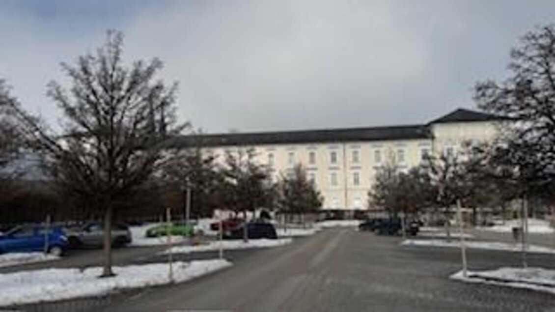 Ein großer, weißer Gebäudekomplex steht am Ende einer schneebedeckten Zufahrt. Rund um das Gebäude sind einige Bäume und Parkplätze sichtbar. | © Hanna Giraudo