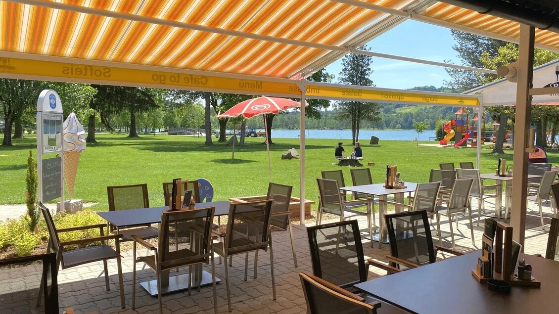 Ein einladendes Restaurant mit Tischen und Stühlen unter einem Sonnenschirm. Im Hintergrund sieht man einen grünen Park und einen ruhigen See. | ©  Familie Stelzer