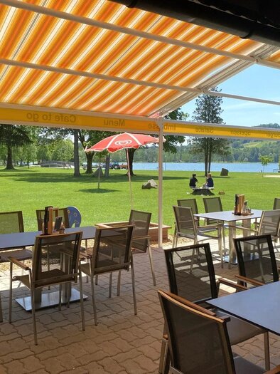 Ein einladendes Restaurant mit Tischen und Stühlen unter einem Sonnenschirm. Im Hintergrund sieht man einen grünen Park und einen ruhigen See. | ©  Familie Stelzer