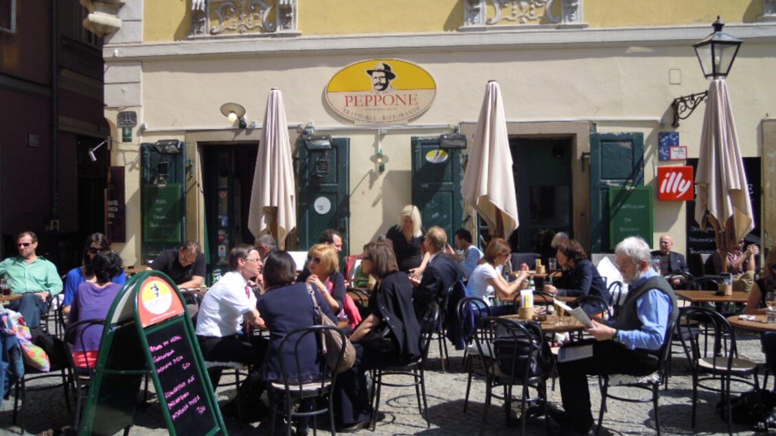 Ein belebtes Café mit vielen Gästen, die im Freien sitzen. Sonnenschirme spenden Schatten an einem klaren Tag. | © CMS