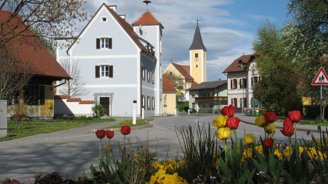 Schöne Straßenansicht mit bunten Blumen im Vordergrund. Im Hintergrund sind traditionelle Häuser und ein Kirchturm sichtbar. | © Schuchlenz