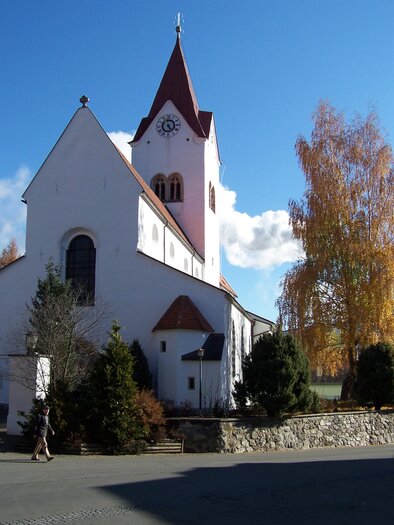 Eine weiße Kirche mit einem roten Dach und einem hohen Turm. Im Hintergrund stehen bunte Bäume und der Himmel ist klar und blau. | © Kth. Kirche Hl. Nikolaus Pöls