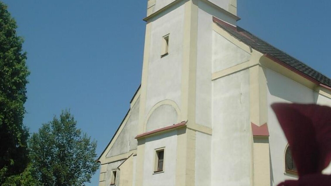 Eine helle Kirche mit einem hohen Turm und einem klaren blauen Himmel im Hintergrund. Rosenblüten sind im Vordergrund sichtbar. | © Marktgemeinde Straß in Steiermark
