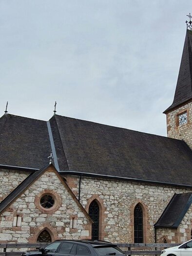 Eine alte steinerne Kirche mit einem hohen Turm. Umgeben von Bäumen und einigen Autos im Vordergrund. | © Stephanie Bor